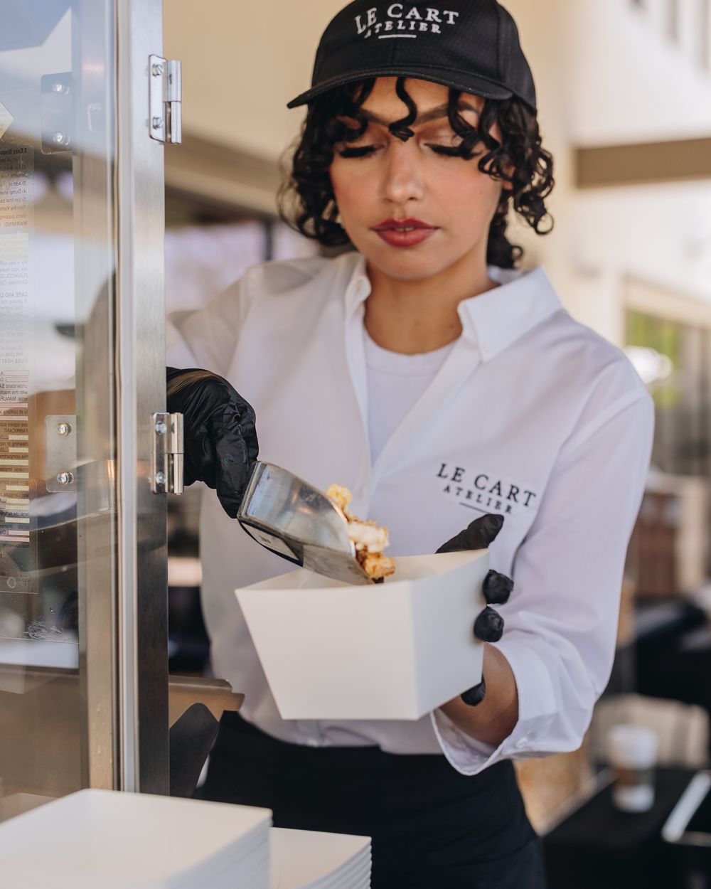 A Le Cart Atelier attendant in a black cap and white shirt scooping fresh popcorn into a box