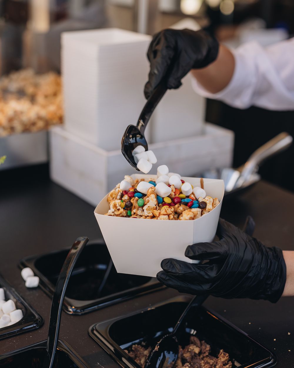 Gloved hands adding marshmallows to a box of caramel popcorn with candy and pretzels